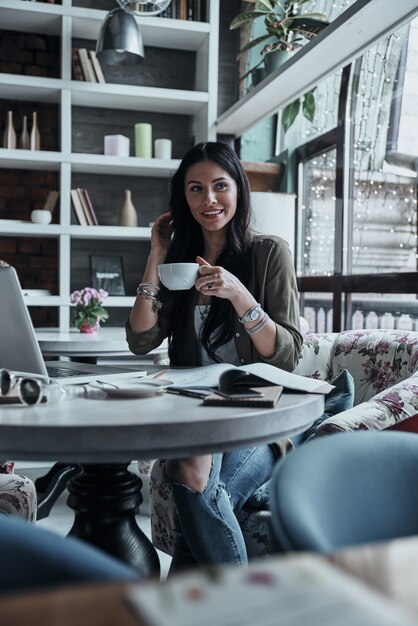 Genieten van verse koffie. Aantrekkelijke jonge glimlachende vrouw die een kopje koffie vasthoudt en wegkijkt