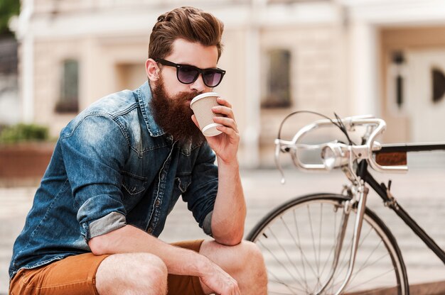 Genieten van verse koffie. Zelfverzekerde jonge bebaarde man die koffie drinkt en naar de camera kijkt terwijl hij buiten in de buurt van zijn fiets zit