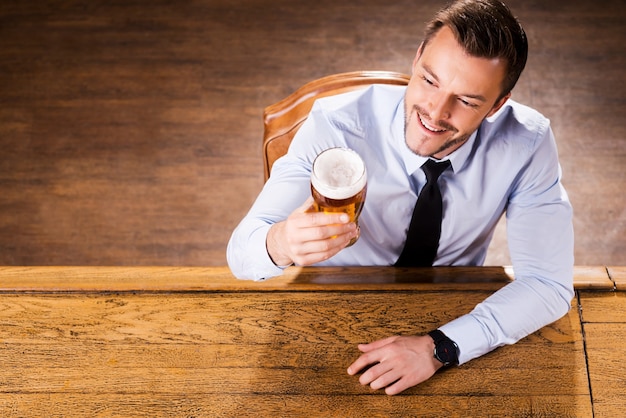 Genieten van zijn favoriete bier. bovenaanzicht van een knappe jongeman in overhemd en stropdas die glas met bier onderzoekt en glimlacht terwijl hij aan de bar zit