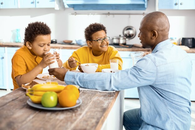 優しいケア。魅力的な若い父親が息子にミルクを一杯与え、テーブルに一緒に座って朝食を食べている間、息子が飲むのを手伝っています