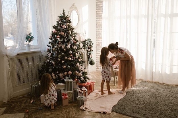 Gentle mother talks with her little daughter while her second little daughter sits next to the gifts under the New Year's tree in the full of light cozy room .