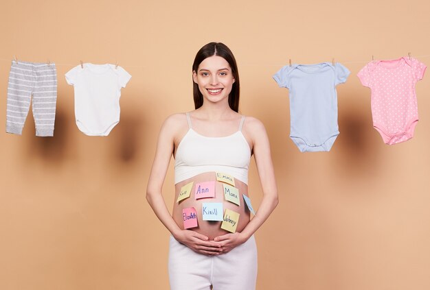 A gentle, sweet, smiling pregnant woman, with a naked belly, with variants of baby names on paper stickers on her tummy, stands on a beige background, preparing for childbirth.