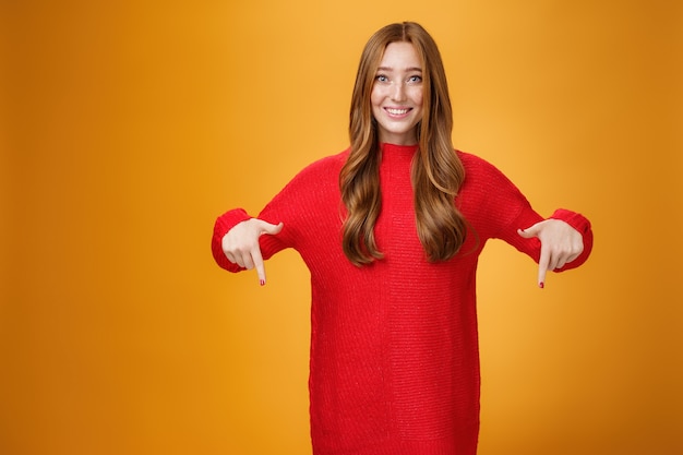 Gentle and tender cute ginger girl with freckles and blue eyes feeling touched and pleased, pointing at heartwarming promotion, showing direction down and smiling against orange background