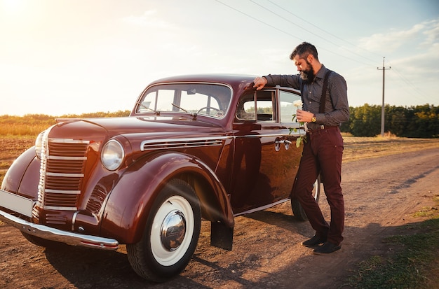 Gentleman met baard in een shirt, broek met bretels houdt een witte roos in zijn hand in de buurt van een bruine retro auto op een landweg en kijkt op zijn horloge