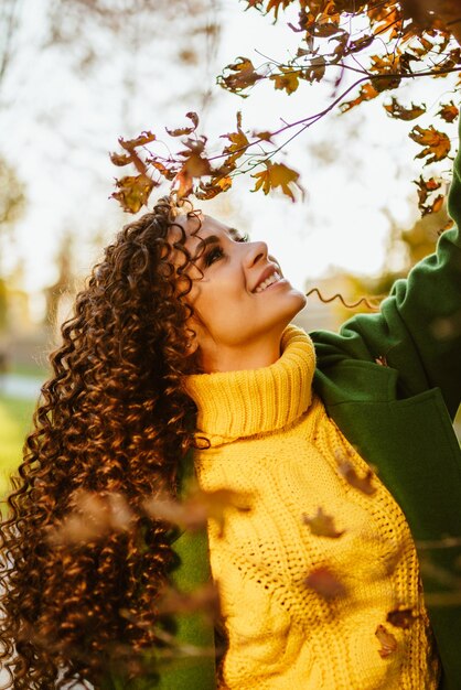 Gently smiling looks at the yellow leaves on the tree lifting up the head curly brunette in a bright yellow sweater