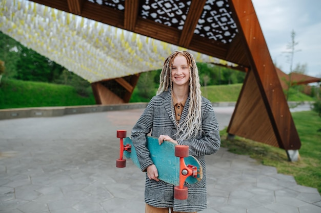 Photo gently smiling teenage girl with dreads holding a skate in a park