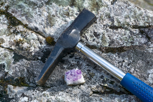 Geological hammer and a sample of amethyst mineral lie on a natural lichen covered rock closeup