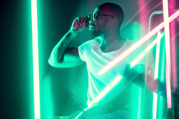 Geometric lines. Cinematic portrait of stylish young man in neon lighted room. Bright neoned colors. African-american model, musician indoors. Youth culture in party, festival style and music concept.