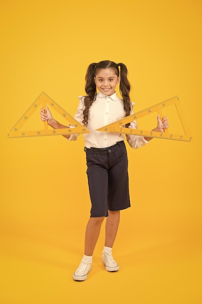 Geometry and measure cute schoolgirl holding triangles for\
geometry lesson on yellow background small child with geometrical\
tools for geometry little girl learning geometry at school