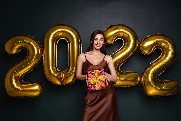 Georgeous brunette lady in beautiful dress holding gift box on black studio background with golden  ...