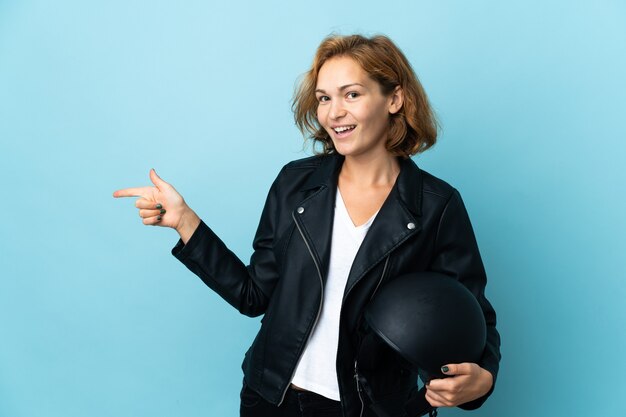 Georgisch meisje dat een motorhelm houdt die op blauwe achtergrond wordt geïsoleerd die vinger aan de kant richt