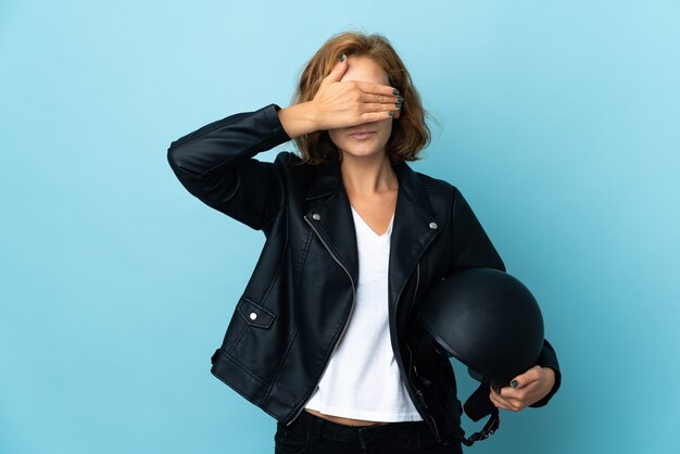 Georgisch meisje dat een motorhelm houdt die op blauwe muur wordt geïsoleerd die ogen behandelt door handen. ik wil niets zien