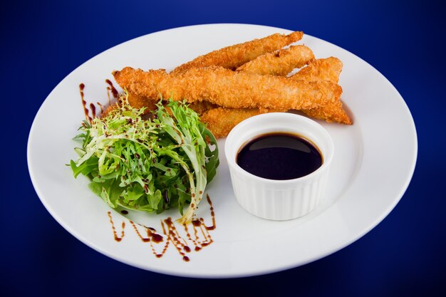 Gepaneerde kipreepjes met saus en slablaadjes. Plat lag bovenaanzicht op een witte plaat op een blauwe achtergrond.