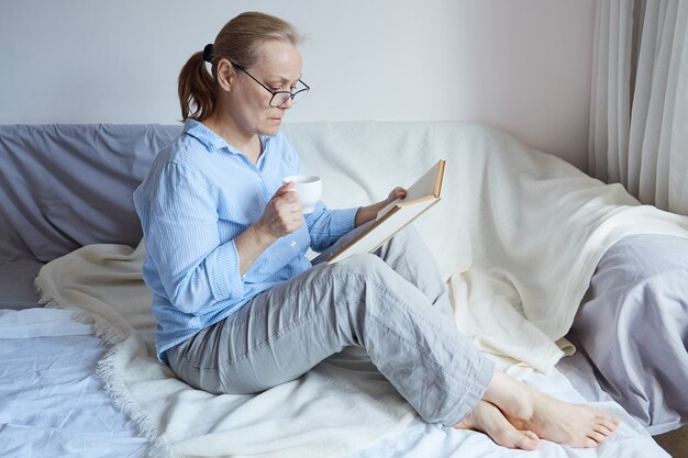 Gepensioneerde m / v vrouw met bril leest een boek, houdt een kopje koffie, zit thuis op de bank.