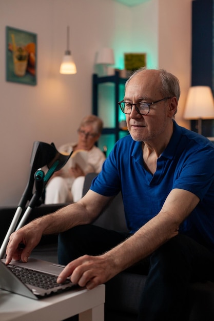 Gepensioneerde oude man die thuis op het toetsenbord van de laptop typt. Kaukasische senior volwassene met apparaat op tafel en oudere vrouw zittend in een rolstoel op de achtergrond. Bejaard paar samen in woonkamer