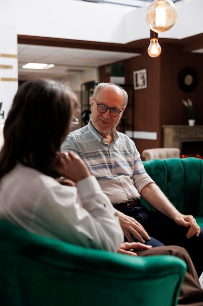 Gepensioneerde oudere echtgenoot en vrouw zitten op comfortabele banken in de lounge en plannen hun vakantieactiviteiten. Gelukkige oudere echtpaar bespreekt vakantieroutine bij exclusieve hotelreceptie.