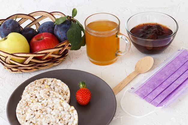 Gepofte rijstwafels, een aardbei op het bord, een kopje thee, een glazen kom met jam, een rieten mand met fruit en een medisch beschermend masker op de witte gestructureerde achtergrond.