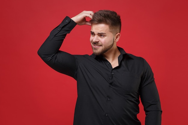 Gepreoccupeerde jonge bebaarde man 20s in klassieke zwarte shirt poseren geïsoleerd op rode muur achtergrond studio portret. Mensen emoties levensstijl concept. Bespotten kopie ruimte. Leg de hand op het hoofd en kijk opzij.