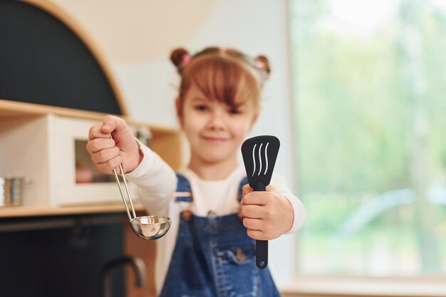 Gereedschap in handen houden en laten zien Klein meisje in vrijetijdskleding heeft plezier door met speelgoed in de keuken te spelen