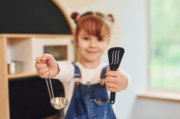 Gereedschap in handen houden en laten zien Klein meisje in vrijetijdskleding heeft plezier door met speelgoed in de keuken te spelen