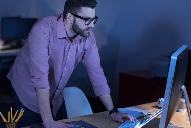 Gericht op werk. Aantrekkelijke slimme bebaarde man op een knop te drukken en te kijken naar het computerscherm terwijl hij ervoor staat