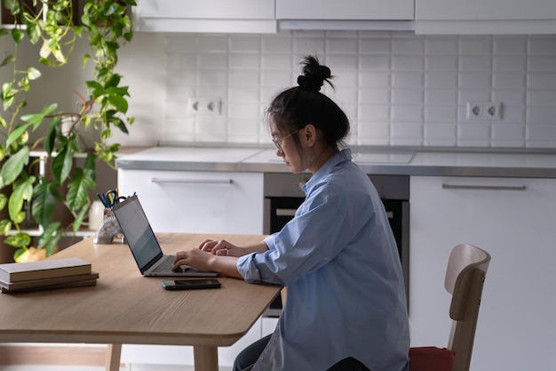 Gerichte Aziatische freelancer die op afstand werkt, zit aan tafel met laptop in de keuken