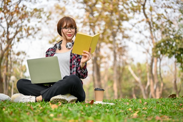 Gerichte Aziatische studente zit op het gras, leest een boek en gebruikt haar laptop