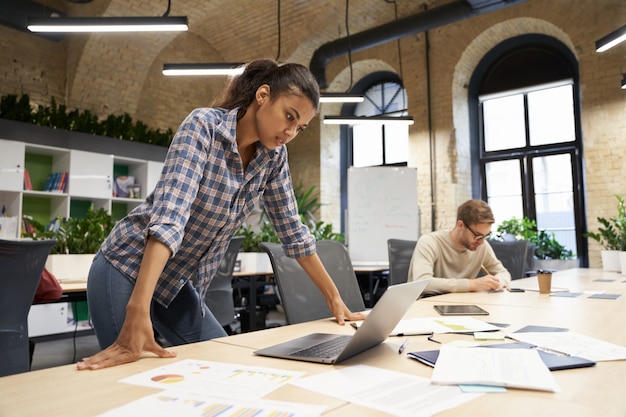 Gerichte jonge zakenvrouw van gemengd ras die in de moderne coworking-ruimte staat en naar laptop kijkt