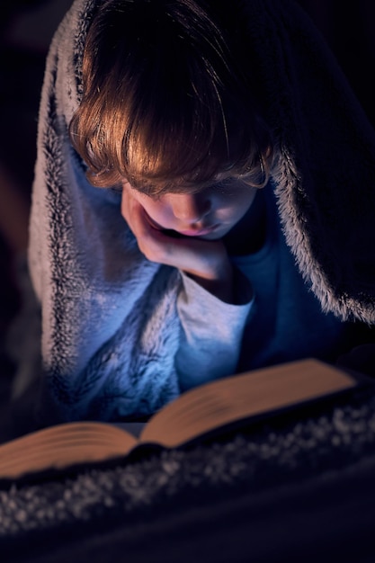 Gerichte jongen gewikkeld in een warme deken die bij de hand leest en een interessant boek leest terwijl hij op bed ligt in de donkere kamer