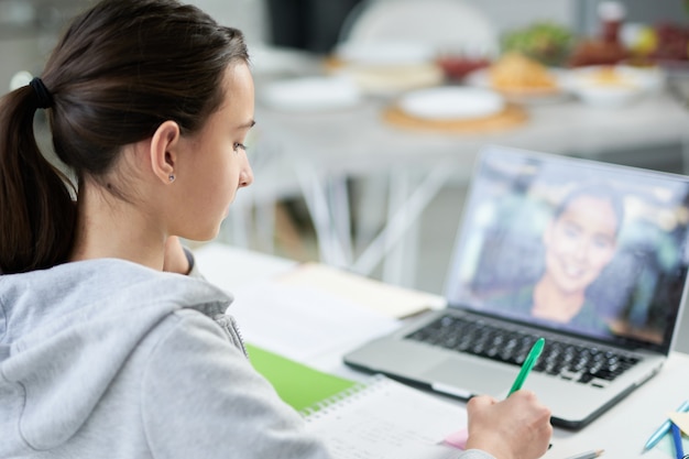 Gerichte Latijns-tiener die in haar schrijfboek schrijft terwijl ze online les heeft, laptop gebruikt, thuis aan tafel zit. Afstandsonderwijs, thuisonderwijs concept