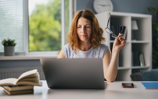 Gerichte rijpe dame in vrijetijdskleding en bril die op moderne laptop werkt terwijl ze in een lichte woonkamer zit Concept van mensentechnologie en werken op afstand