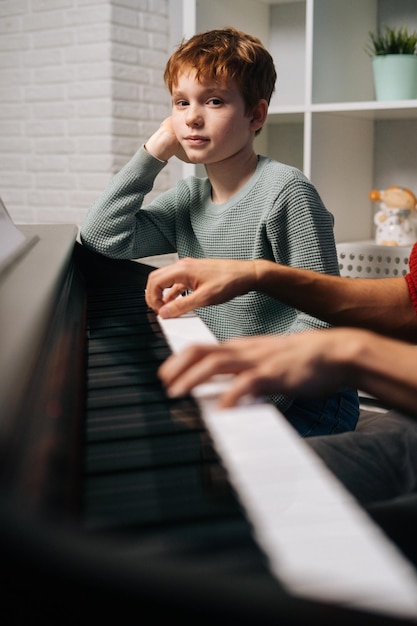 Gerichte roodharige kindstudent die zijn leraar luistert terwijl hij piano speelt tijdens de les en naar de camera kijkt