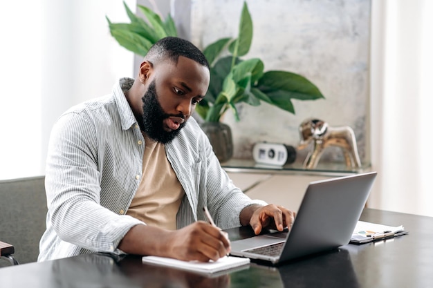 Gerichte succesvolle slimme Afro-Amerikaanse mannelijke bedrijfseigenaar die aan een bureau zit in een modern kantoor en aan een project werkt met documenten die het maken van aantekeningen analyseren, bereidt een rapport voor