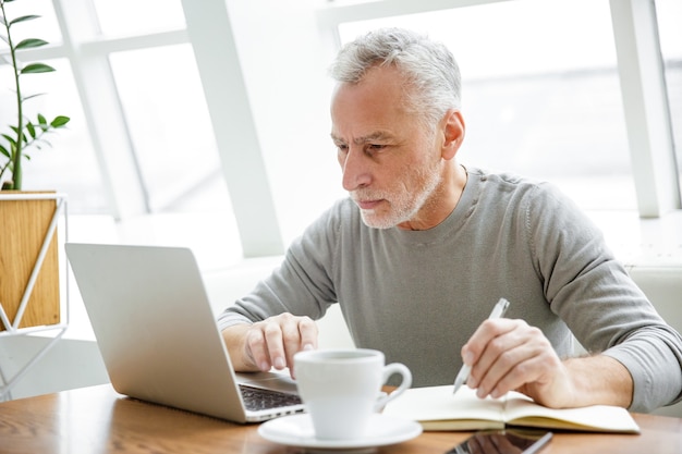 gerichte volwassen man die aantekeningen maakt en met laptop werkt terwijl hij binnenshuis in café zit