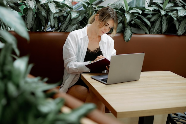 Gerichte vrouwelijke schrijfnotities in dagboek op bruine bank aan cafétafel Lady freelancen met laptop omgeven door planten