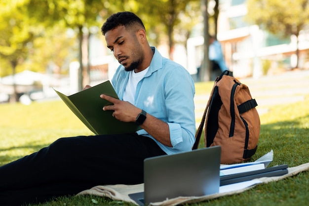 Gerichte zwarte mannelijke student die met behulp van laptop leert en aantekeningen maakt terwijl hij buiten in het park zit