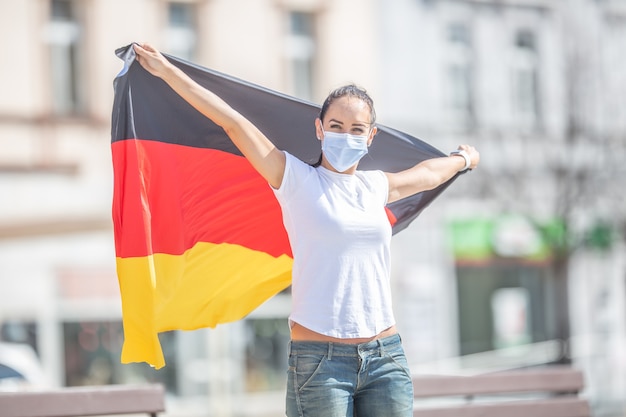 German fan in a face mask holds the flag behind her outdoors, smiling.