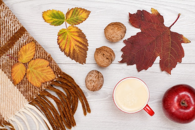 Geruite plaid, droge gele en bruine bladeren, kopje koffie, appel en walnoten op houten ondergrond. Een herfstcompositie. Bovenaanzicht.