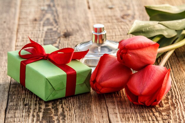 Geschenkdoos omwikkeld met rood lint en fles parfum op houten planken met rode tulpen.