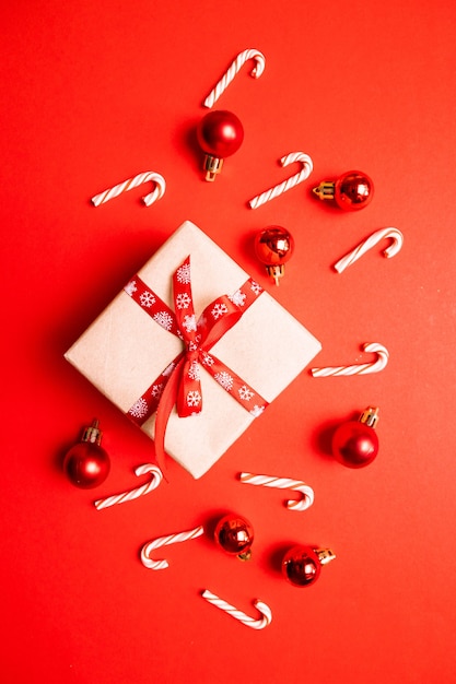 Geschenkdoos verpakt in ambachtelijk papier met rood lint boog, riet van het suikergoed, kerstballen op een rode achtergrond. Moderne creatieve minimale vakantiesamenstelling in effen kleur