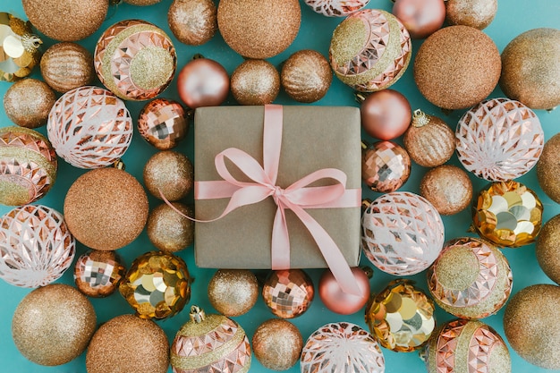 Geschenkdoos verpakt in ambachtelijk papier met roze lint en gouden kerstboomballen op lichtblauwe achtergrond met glitters.