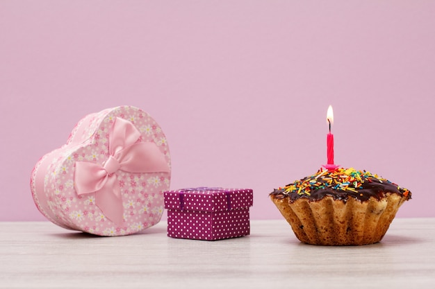 Geschenkdozen en smakelijke verjaardagsmuffin met chocoladeglazuur en karamel, versierd met brandende feestelijke kaars op lila achtergrond. Gelukkige verjaardag minimaal concept.