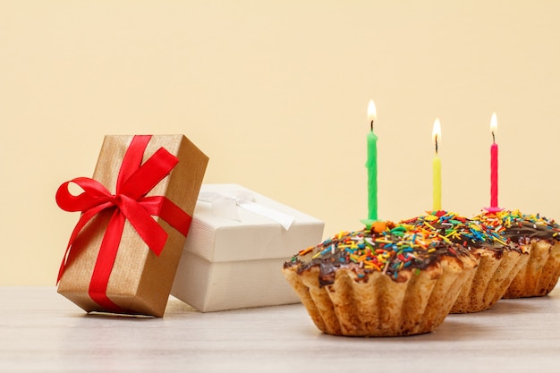 Geschenkdozen en smakelijke verjaardagsmuffins met chocoladeglazuur en karamel, versierd met brandende feestelijke kaarsen op houten en beige achtergrond. Gelukkig verjaardagsconcept.