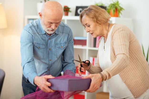 Foto geschenkverpakkingsproces. vrolijke oude vrouw die een schaar in haar handen houdt terwijl ze haar vriend helpt met het inpakken van cadeaus