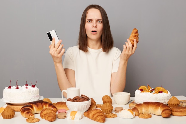 Geschokt, bang, vrouw, vervelend, witte, t-shirt, geïsoleerde, op, grijze achtergrond, zitten aan tafel, met, desserts, vasthouden, gsm, en, croissant, kijken naar van fototoestel, met, bang, uitdrukking