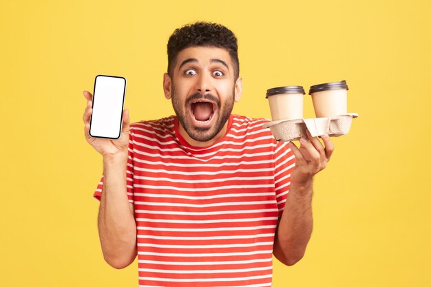 Geschokt bebaarde man in gestreept t-shirt schreeuwend in handen witte display smartphone en twee papieren bekers met koffie verrast met snelle levering. Indoor studio-opname geïsoleerd op gele achtergrond