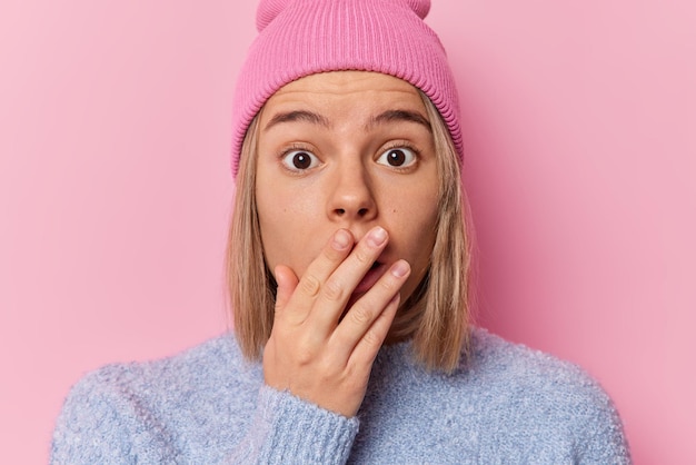 Geschokt beschaamde jonge vrouw heeft kort haar bedekt mond met hand hoort iets onverwachts en adembenemend draagt hoed en casual zachte jumper poses tegen roze achtergrond. Omg-concept.