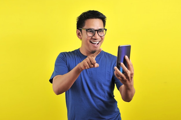 Geschokt gezicht van Aziatische man in blauw shirt kijkend naar telefoonscherm op gele achtergrond.