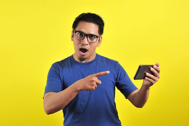 Geschokt gezicht van aziatische man in blauw shirt kijkend naar telefoonscherm op gele achtergrond.