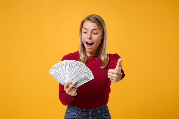 Geschokt jong blond vrouwenmeisje in vrijetijdskleding poseren geïsoleerd op geeloranje achtergrond. Mensen levensstijl concept. Bespotten van kopieerruimte Houd fan van contant geld in dollarbiljetten met duim omhoog.